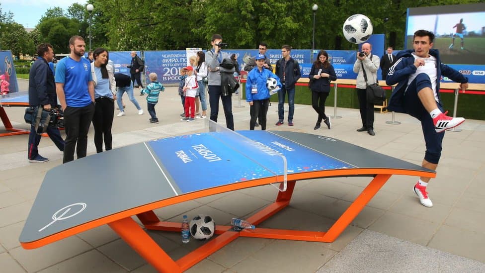 Niños y adultos participando en un reto de fútbol en una plaza festiva.